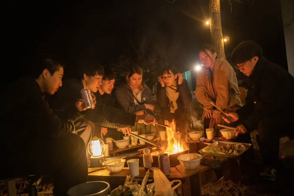 夜の校庭でランタンと焚き火を囲み、鍋を分け合う人たち